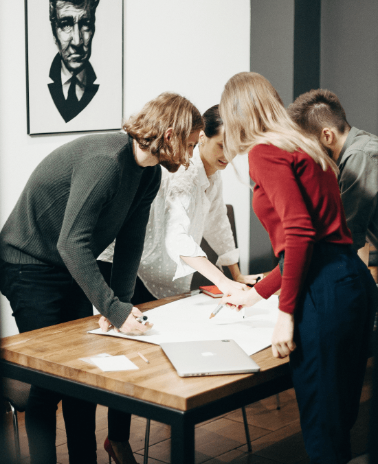 Four people with markers work together on a large sheet of paper on a table.