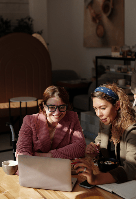 Two women look at a laptop on a table in a coffeeshop.