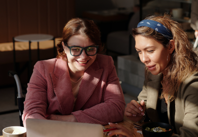Two women look at a laptop on a table in a coffeeshop.