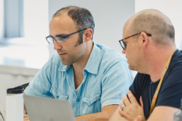 Two men with laptops sit in a classroom, talking.